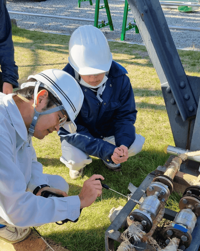 Two inspectors in helmets crouching at an outdoor steam trap manifold, holding a waveguide against the housing